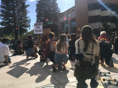 A group of peaceful protesters, mostly female, mostly white, kneeling with their backs to the camera. A woman of color holds a sign "Defund the Police"